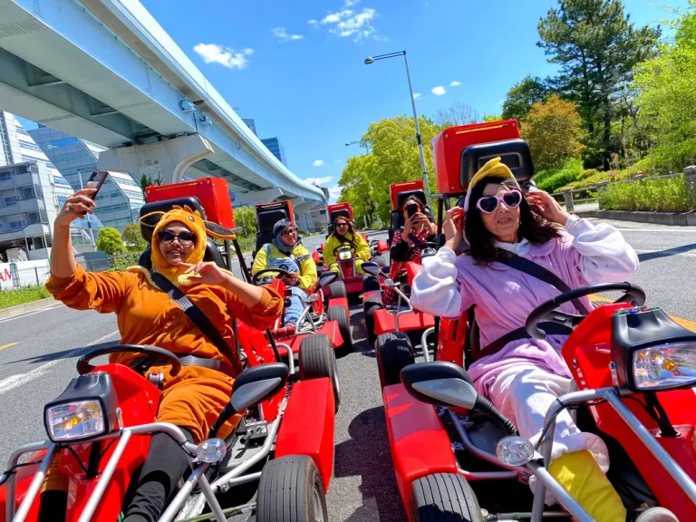 Group of friends in colorful animal-costume outfits ride red go-karts down a sunny city street, posing for selfies while others follow in the background under a blue sky.