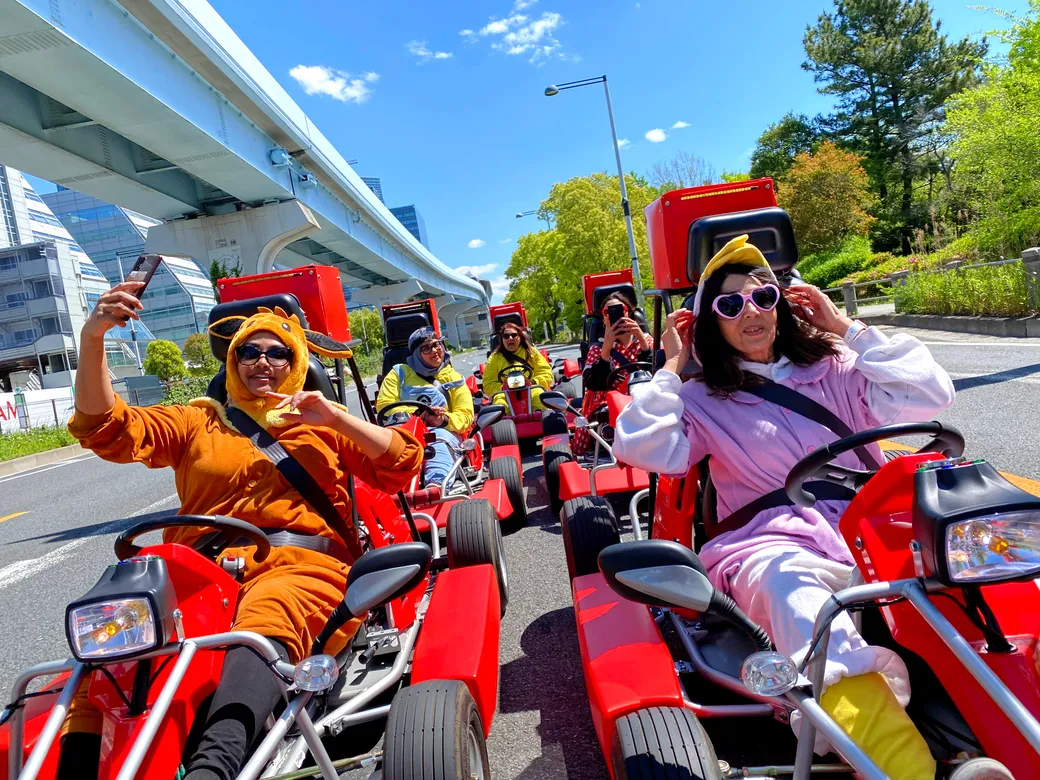People in colorful costumes riding red go-kart style scooters on a city street, posing for selfies under a blue sky, with a person in orange animal onesie taking a selfie.