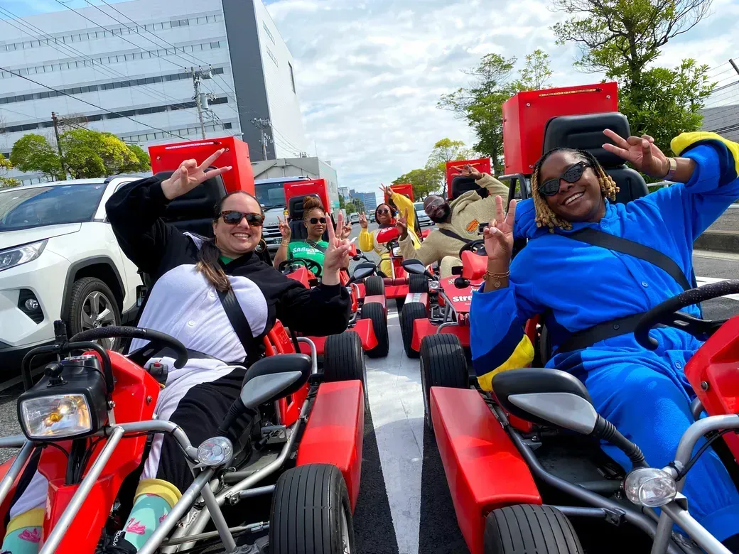 Group of people in red go-karts on a city street, smiling and flashing peace signs at the camera with a blue sky above.