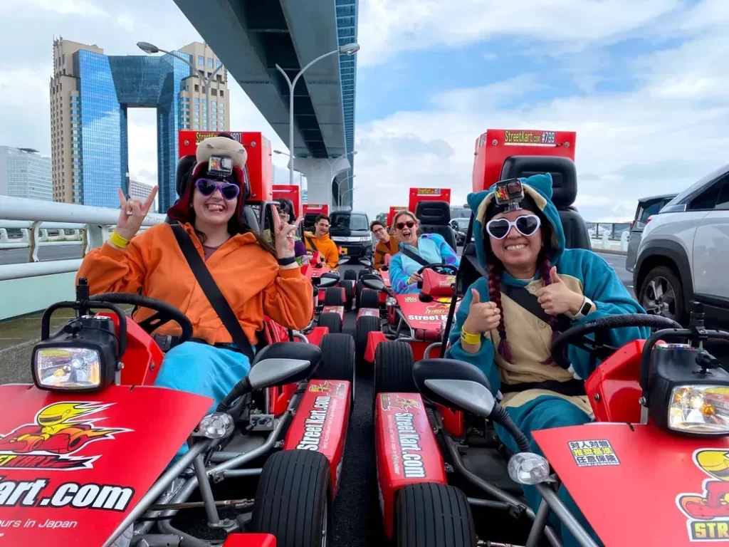 Group of friends in colorful outfits in red go-karts, giving thumbs up on a street go-kart ride.