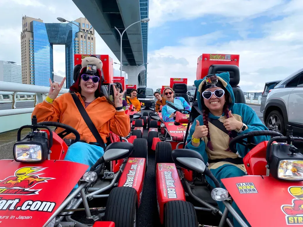 Group of friends in colorful outfits in red go-karts, giving thumbs up on a street go-kart ride.